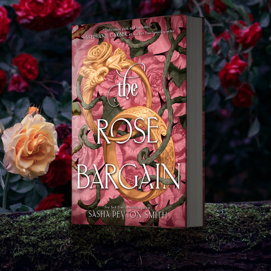 A hardcover copy of THE ROSE BARGAIN by Sasha Peyton Smith atop a mossy log. The book cover features pink roses, green vines, and 2 golden wedding bands.