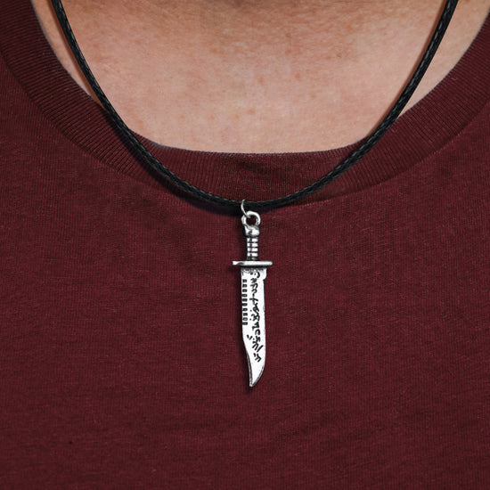 Close up of a necklace pendant against a model's maroon shirt. The pendant is shaped like a hunting knife with mysterious symbols carved into the blade. A black braided cord is attached to the hilt.