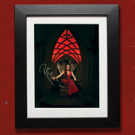 A picture in a black frame against a dark red wall. The picture is of Ruth Connell as the character "Rowena" from the TV series Supernatural, sitting on her throne. Above her is a red stained glass window with an upside down cross at the top. Next to her is Ruth's signature in white ink.