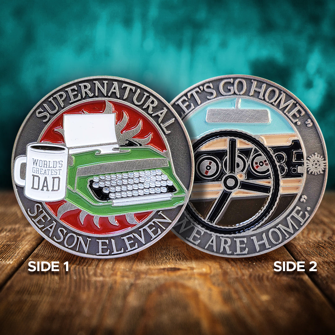 Front and back views of a brass coin. On the front is a green typewriter on a red background. Next to it is a white mug with "World's Greatest Dad" printed on it. Around the edge of the coin, raised text says "Supernatural Season Eleven." The back of the coin depicts a black steering wheel in front of a car dashboard. Around the edge is raised text saying "Let's go home... we are home."