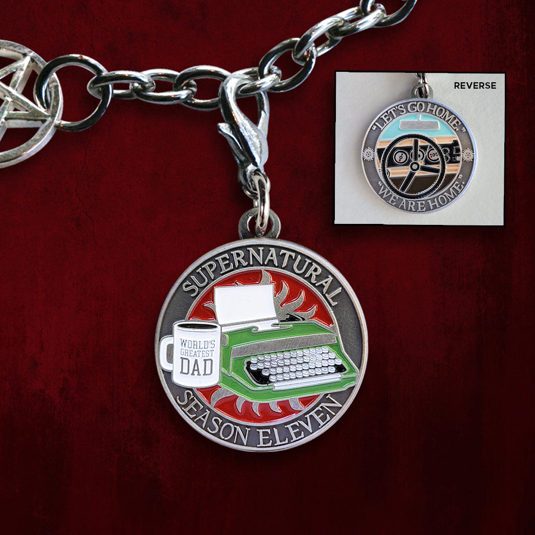 Front and back views of a brass charm. On the front is a green typewriter on a red background. Next to it is a white mug with "World's Greatest Dad" printed on it. Around the edge of the coin, raised text says "Supernatural Season Eleven." The back of the coin depicts a black steering wheel in front of a car dashboard. Around the edge is raised text saying "Let's go home... we are home." Behind the charm is a red background.