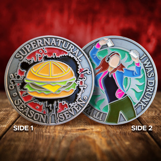 A brass coin charm with "Supernatural season seven" and a burger against a red and black background on one side and "It was comic-con. I was drunk." with teal background, an anti-possession symbol, and a sillhouette of Charlie Bradbury on the other. The charms are on a wood table, in front of a red wall.