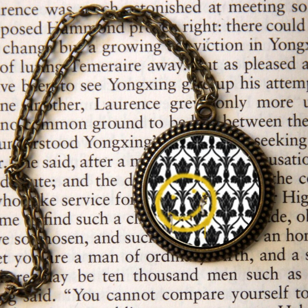 Close up of a round bronze pendant against a page of text. The center of the pendant has a black on white fleur-de-lys wallpaper pattern. On top of the wallpaper is a smiley face painted in yellow. The pendant is attached to a bronze link chain.