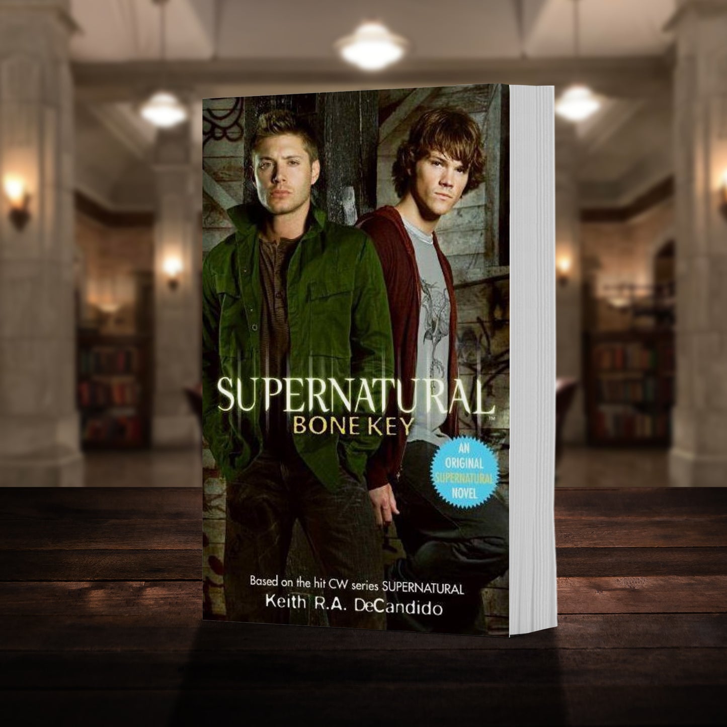 A copy of the book "Supernatural: Bone Key" pictured in the "bunker" from Supernatural. The cover shows early-season Sam and Dean Winchester and reads "Supernatural Bone Key - Based on the hit CW series SUPERNATURAL by Keith R.A. DeCandido"