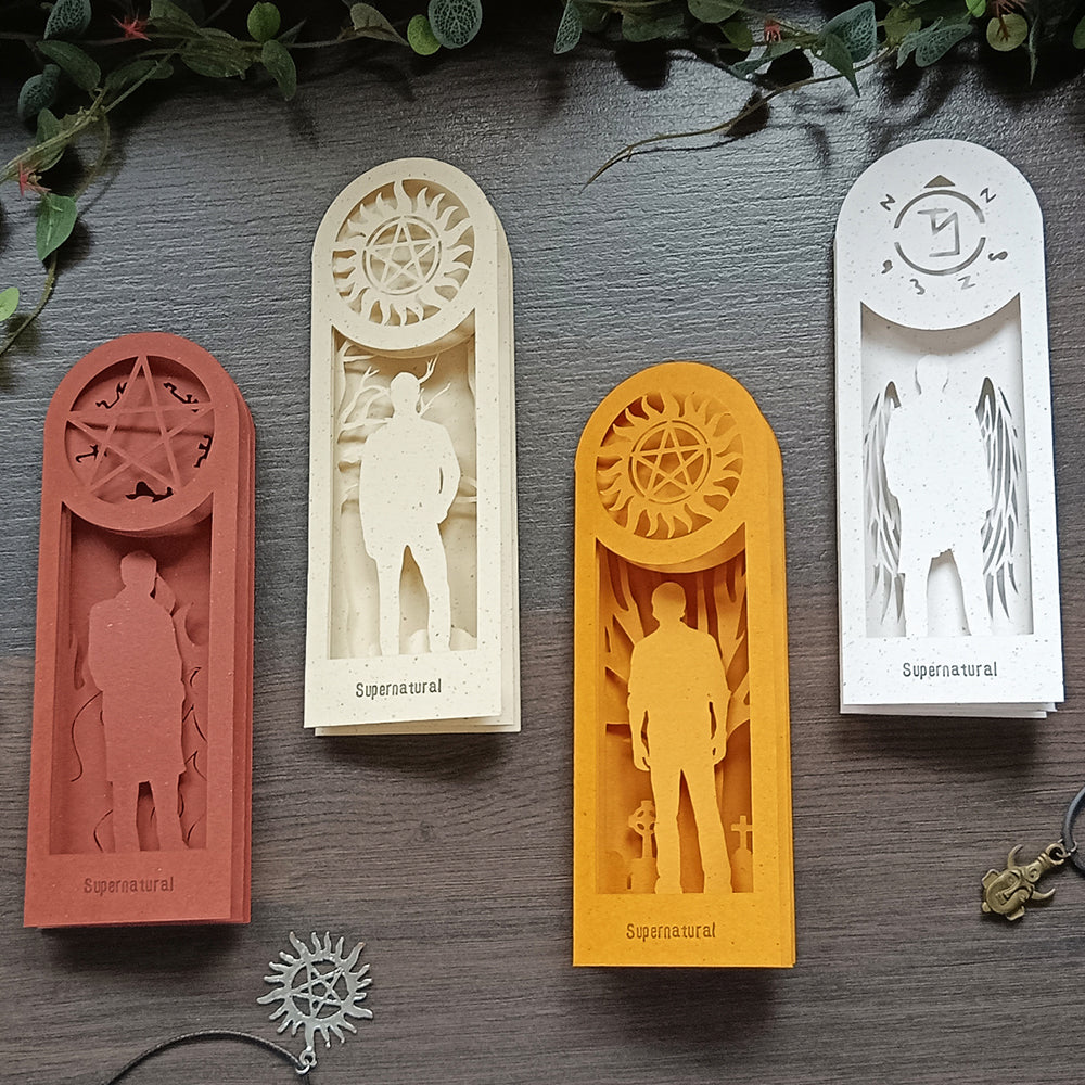 Four hand-carved paper bookmarks, each a different color, on a black tabletop under green leaves. Each bookmark depicts a character from the TV series Supernatural. The red bookmark depicts Crowley. The ivory bookmark depicts Dean Winchester. The yellow bookmark depicts Sam Winchester. The white bookmark depicts Castiel.