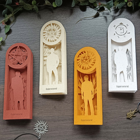 Four hand-carved paper bookmarks, each a different color, on a black tabletop under green leaves. Each bookmark depicts a character from the TV series Supernatural. The red bookmark depicts Crowley. The ivory bookmark depicts Dean Winchester. The yellow bookmark depicts Sam Winchester. The white bookmark depicts Castiel.