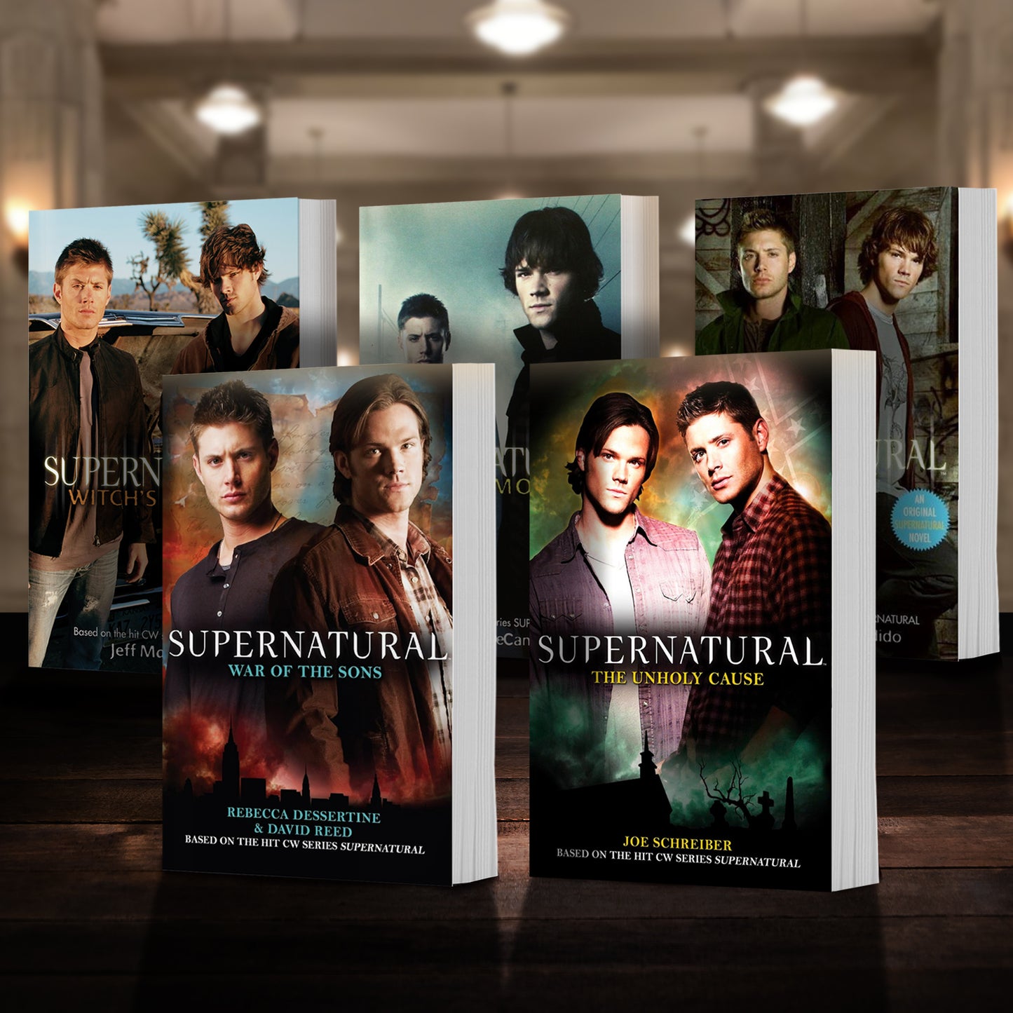 Load image into Gallery viewer, Five novels standing next to each other on a wooden floor. Each book is a novel based on the TV series Supernatural. Behind the books is a library.
