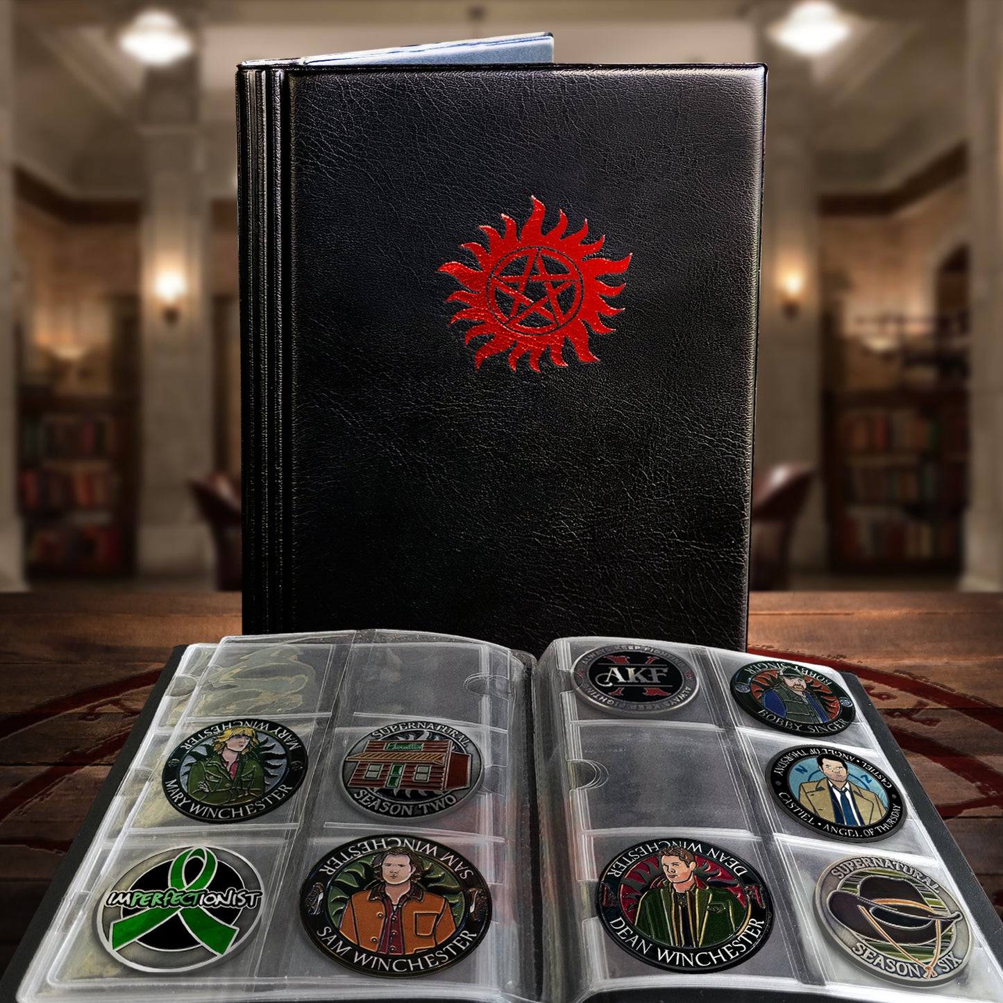 A black faux-leather binder standing upright on a wooden table, with a red anti-possession emblem on the cover. In front of it are two clear plastic coin display pages containing round commemorative challenge coins.