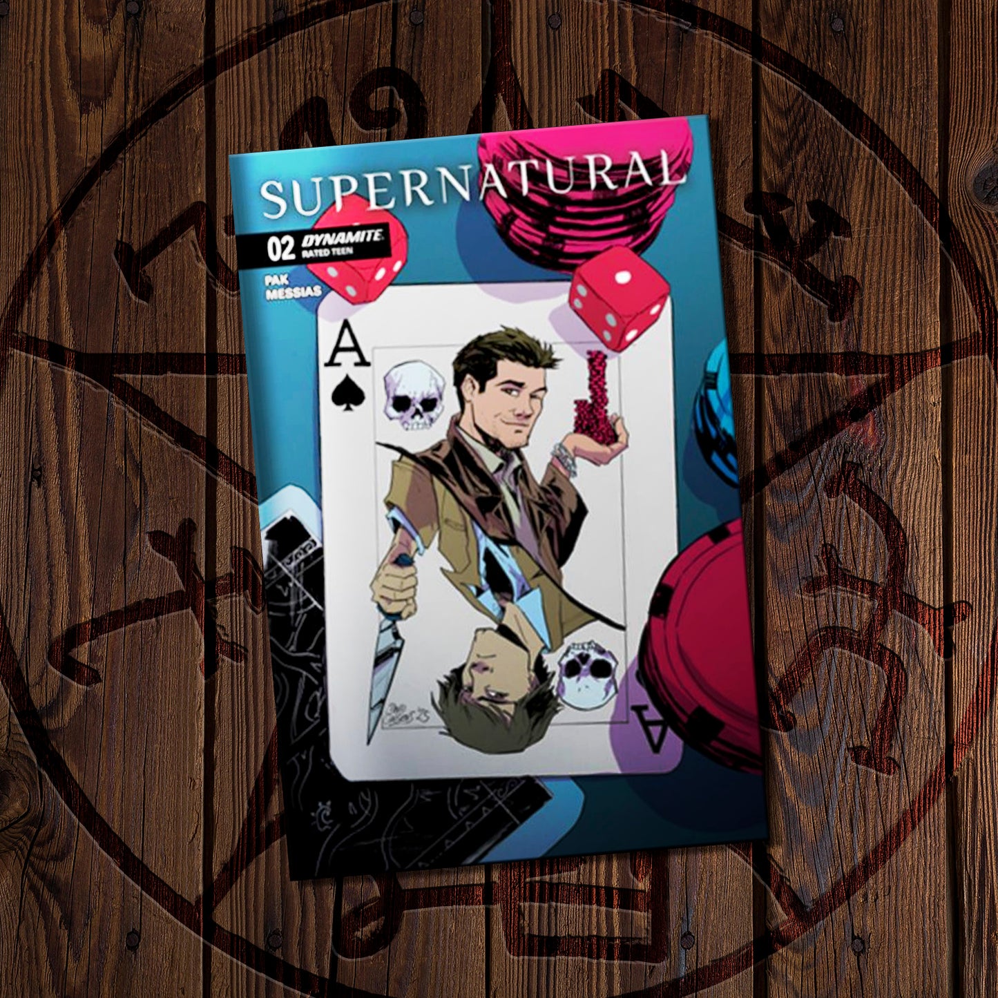 Comic book titled 'Supernatural' on a wooden surface. The cover depicts a drawing os a playing card that features Sam and Castiel, surrounded by dice and poker chips.