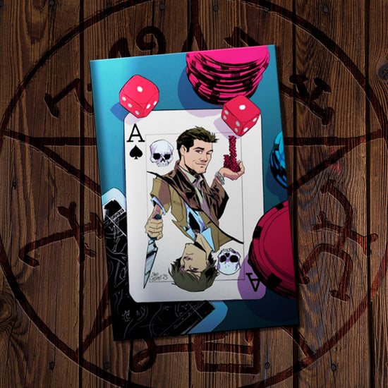 Comic book on a wooden surface. The cover depicts a drawing os a playing card that features Sam and Castiel, surrounded by dice and poker chips.