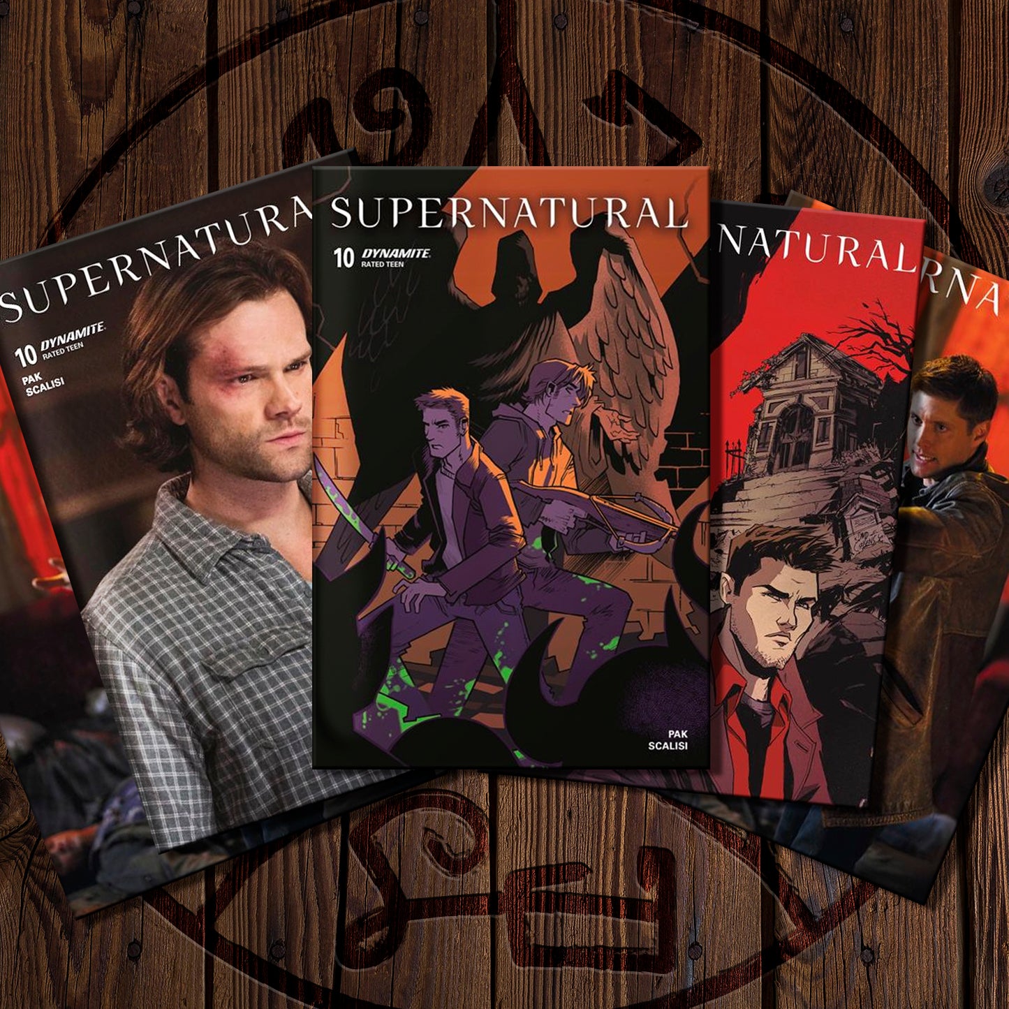 A fan arrangement of three 'Supernatural' comic book volumes on a rustic dark wood background with a circular occult symbol. The covers feature character art and photos of the cast of the show, including Dean Winchester and Sam Winchester.