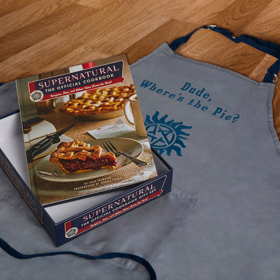 A grey apron on a wood floor. On the apron is blue text saying :dude, where's the pie?" with the anti-possession symbol beneath it. On top of the apron is a blue box, with a cokbook on the box top. The cookbook has a picture of a slice of pie on a plate, with "supernatural, the official cookbook" at the top in white letters.