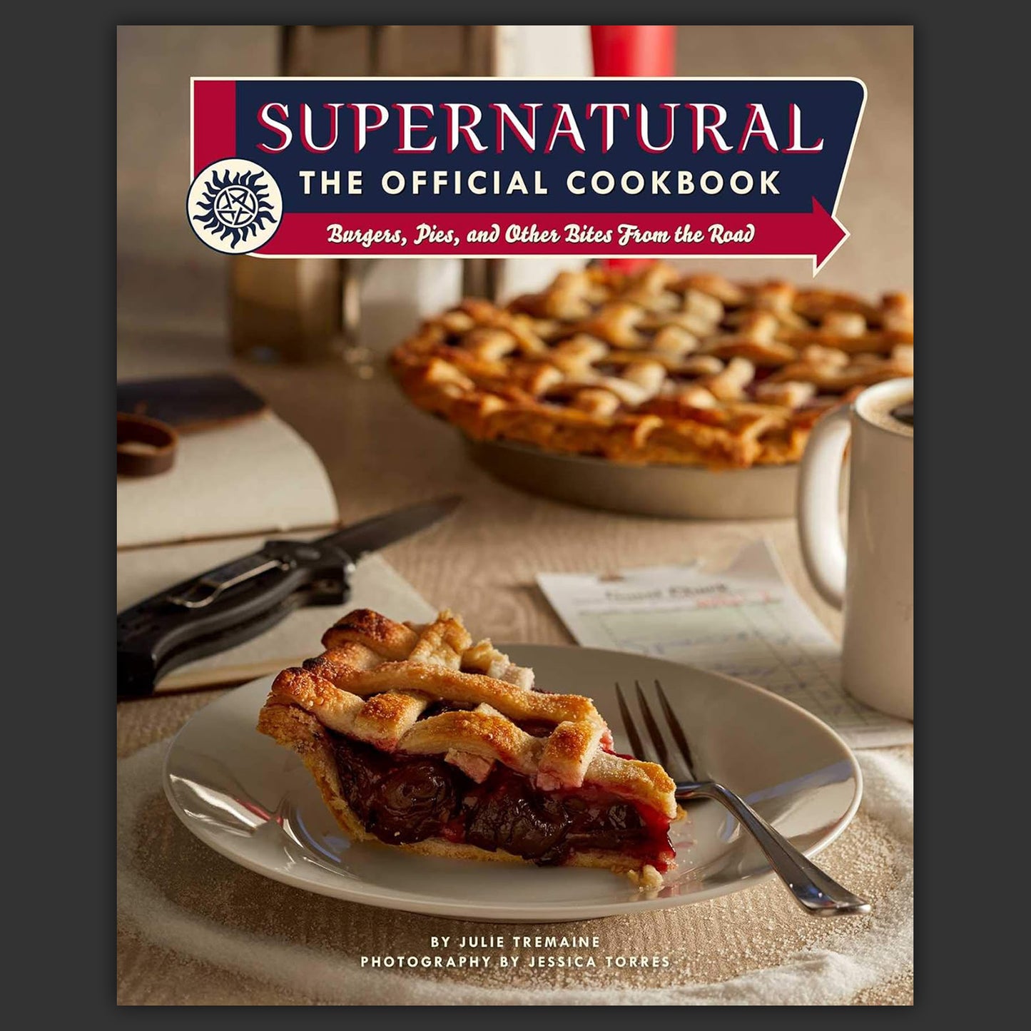 A hardcover cookbook on a white background. The cookbook has a picture of a slice of pie on a plate, with "supernatural, the official cookbook" at the top in white letters.