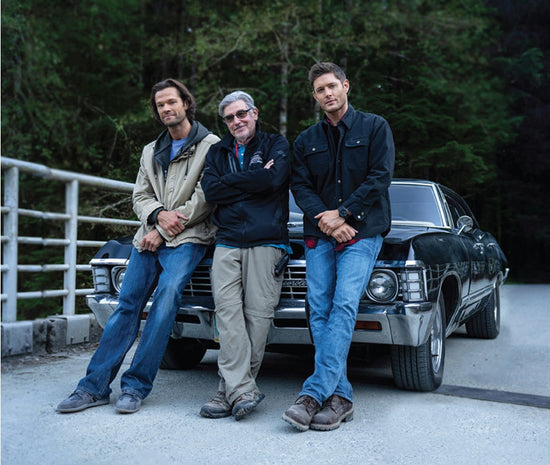 An image of Jensen Ackles and Jared Padalecki with a crew member, standing against the fron of the show's 1967 Impala, parked on a bridge.