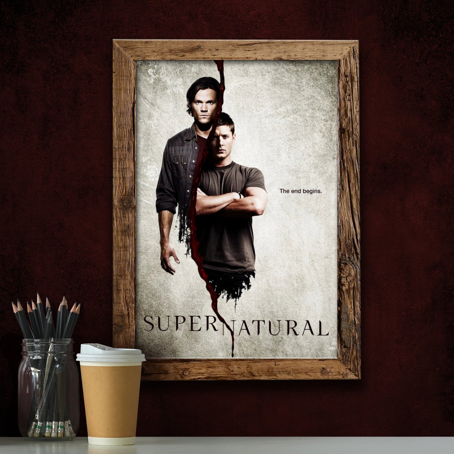 A poster in a wood frame against a dark red background. The poster depicts the characters Sam and Dean Winchester from the TV series "Supernatural," with a bloody line down the center. Text on the right says "The end begins." At the bottom is black text that says "Supernatural" with a bloody line down the center. In front of the poster is a paper coffee cup and a glass jar filled with pencils.