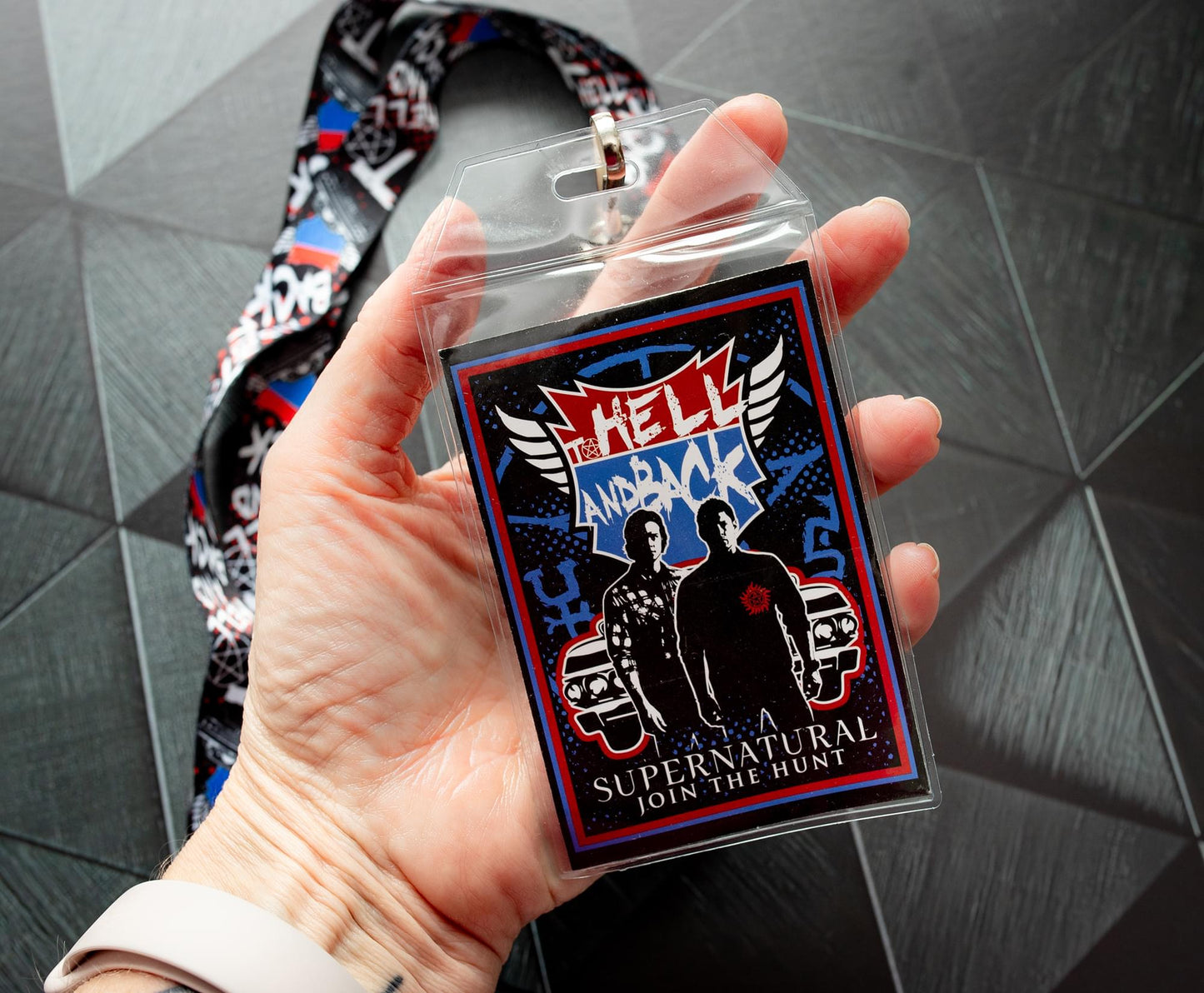 A model's hand holding a lanyard with an anti-possession charm in red and black attached to the end. The lanyard features "to hell and back" in white text. Next to the lanyard is a badge featuring Sam and Dean Winchester. A red and blue road symbol at the top has white text saying to hell and back. The lanyard and badge are above a black surface.