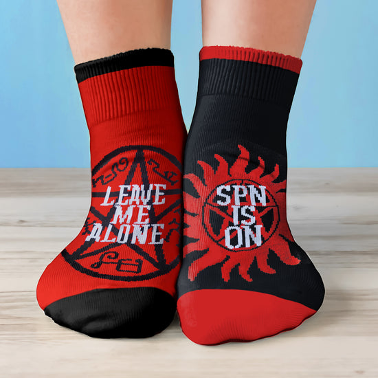 A model's feet wearing black and red socks. One sock has a black top and toes, with a red center, the other is the reverse. The left sock has a black devil's trap symbol on the front, with white text saying "Leave me alone." The right sock has a red anti-possession symbol on the front, with white text saying "SPN in on."