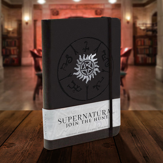 A black hardcover journal on a wooden table, set inside The Bunker. On the front cover is a white anti-possession symbol, set inside a black Devil's Trap symbol. Across the bottom is a white banner, with black text that says "supernatural, join the hunt."