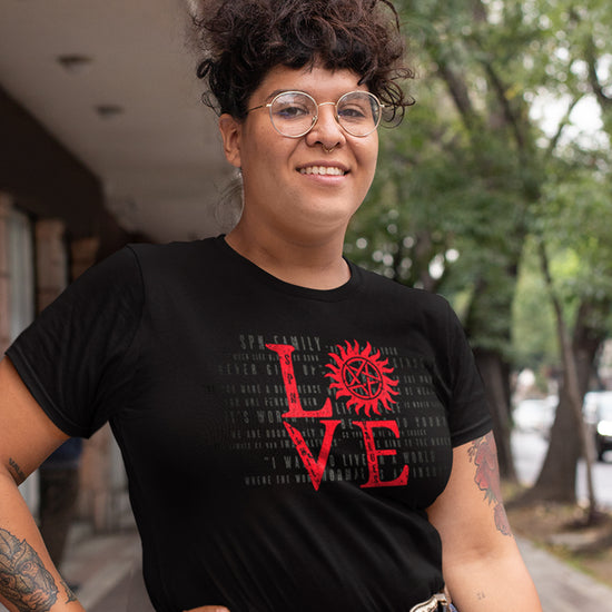 A female model wearing a black unisex t-shirt with red lettering that says LOVE - the "o" is the anti-possession symbol. Behind the model is a street lined with trees.
