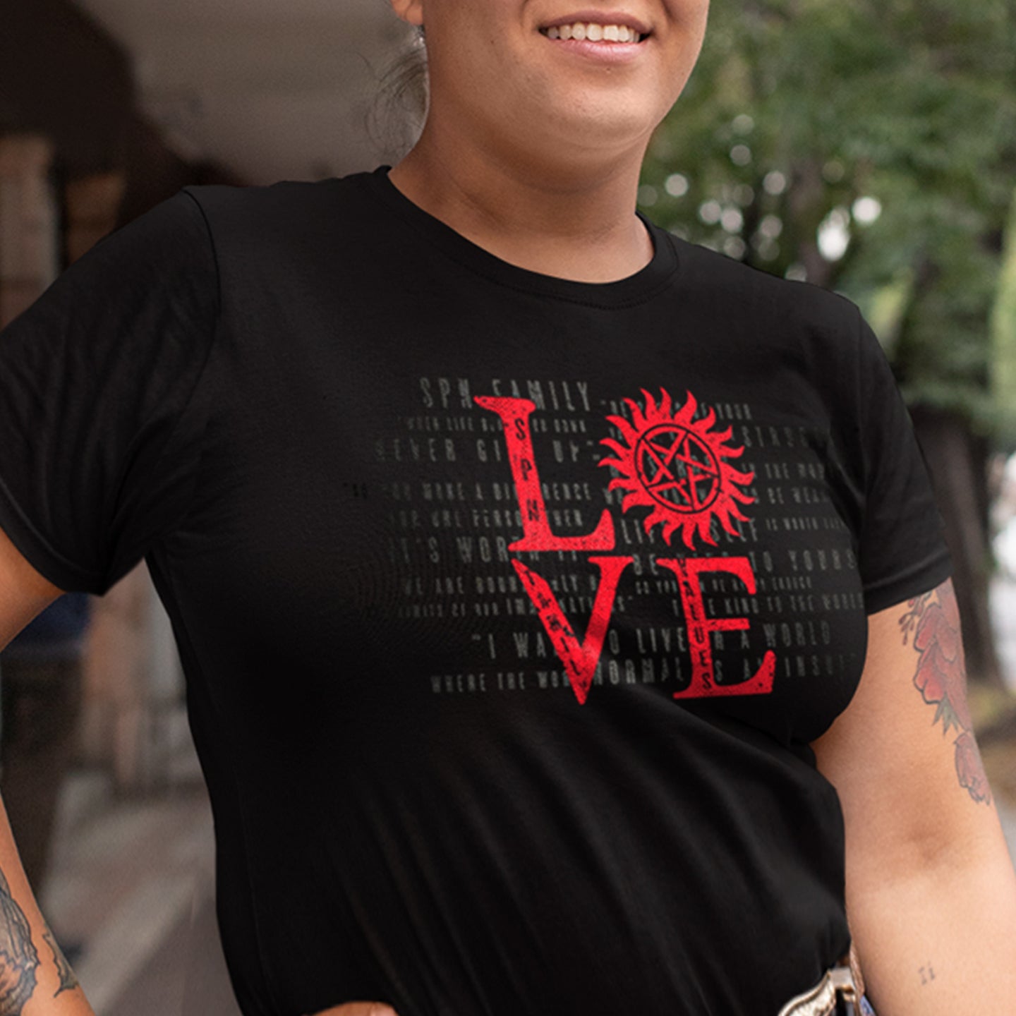 A female model wearing a black unisex t-shirt with red lettering that says LOVE - the "o" is the anti-possession symbol. Behind the model is a street lined with trees.