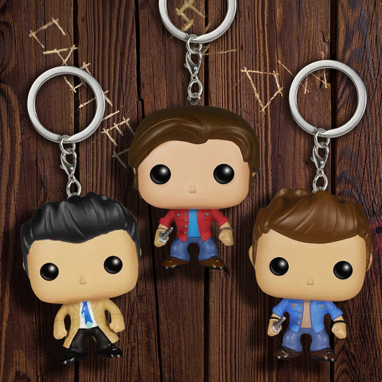 Three keychains side by side against a wooden wall. The keychain in the center is in the shape of Sam Winchester, wearing a red shirt and jeans and carrying a knife. The keychain on the left is in the shape of the angel Castiel, wearing a tan raincoat and blue tie. The keychain on the right is in the shape of Dean Winchester, wearing a blue shirt and carrying a knife.