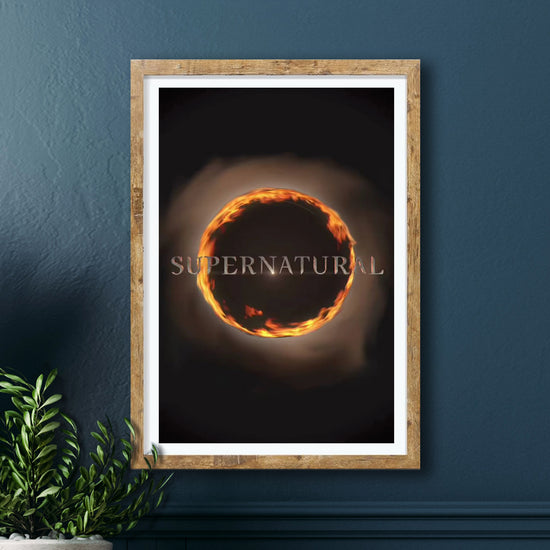 A framed print against a dark blue wall. The print depicts a circle of fire against a black background. Smoke colored text in front of the circle says "supernatural.