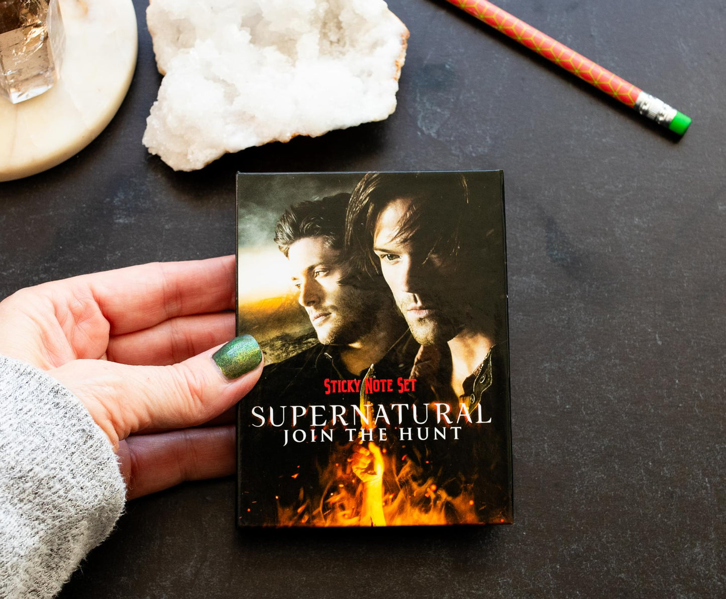 A model's hand holding a sticky note box set featuring Sam and Dean Winchester on the front. Red and white text says Supernatural, Join The Hunt, sticky note set. The set is on a table next to a pencilm salt and a denim jacket.