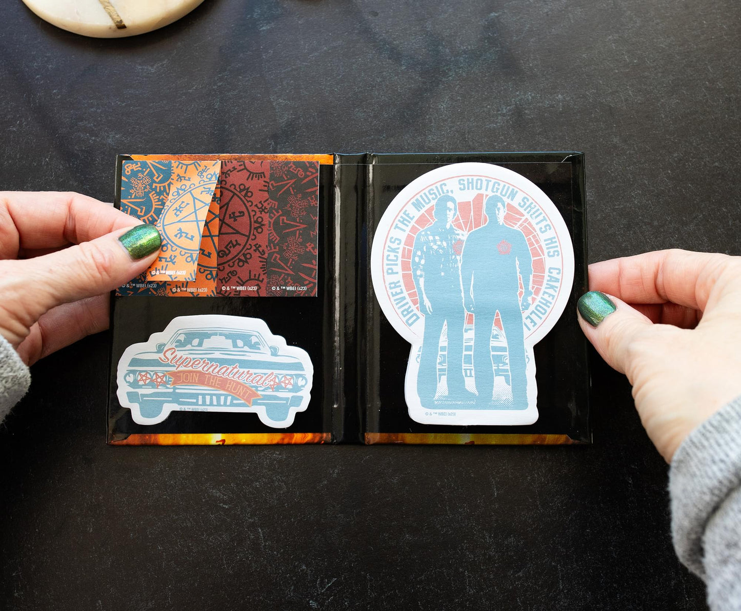 A model's hand holding a collection of Supernatural-themed sticky notes, featuring Sam and Dean, Baby, and the Devils Trap symbol.