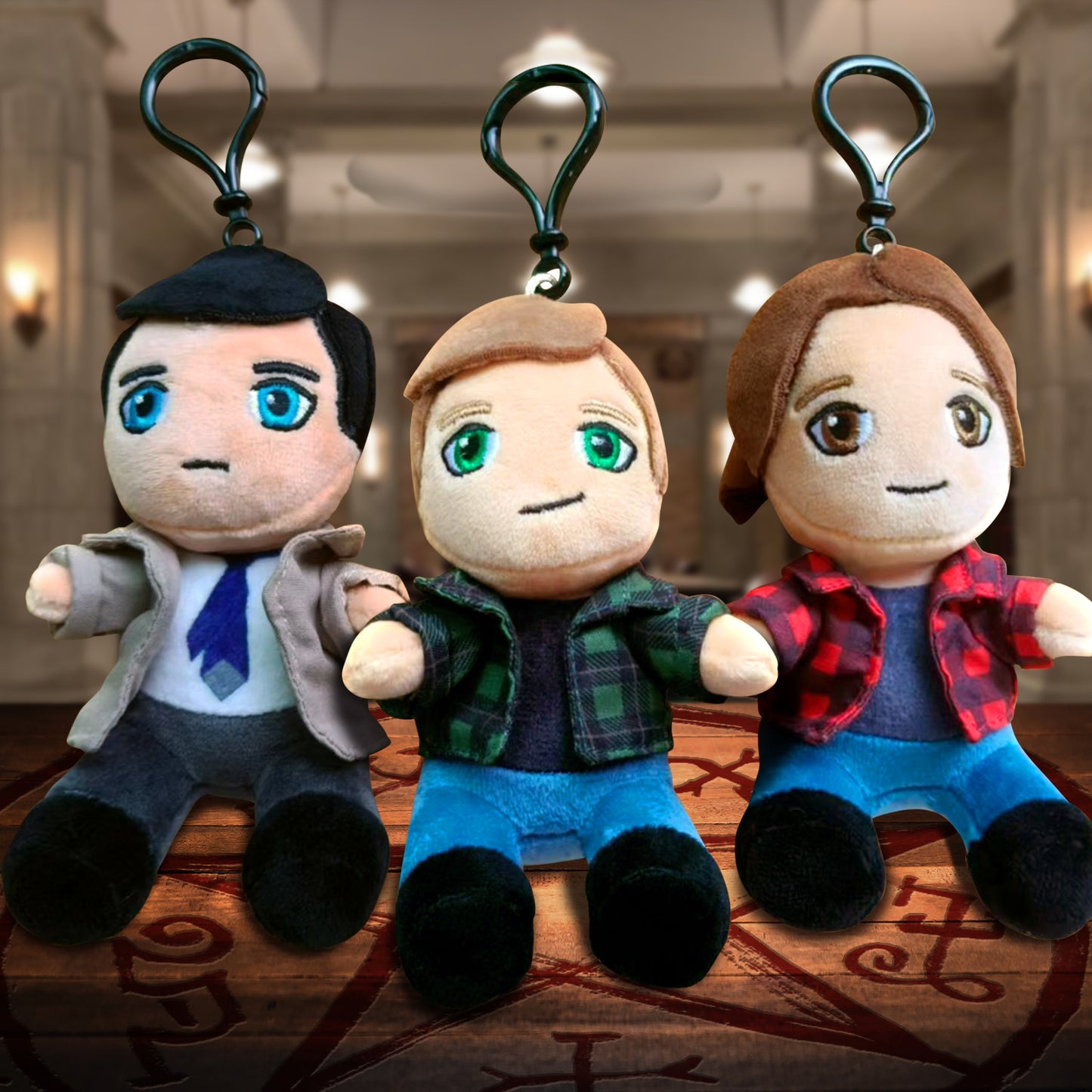 Three plush keychains depicting Castiel, Dean Winchester, and Sam Winchester. Below them is a Devil's Trap symbol on a wooden floor