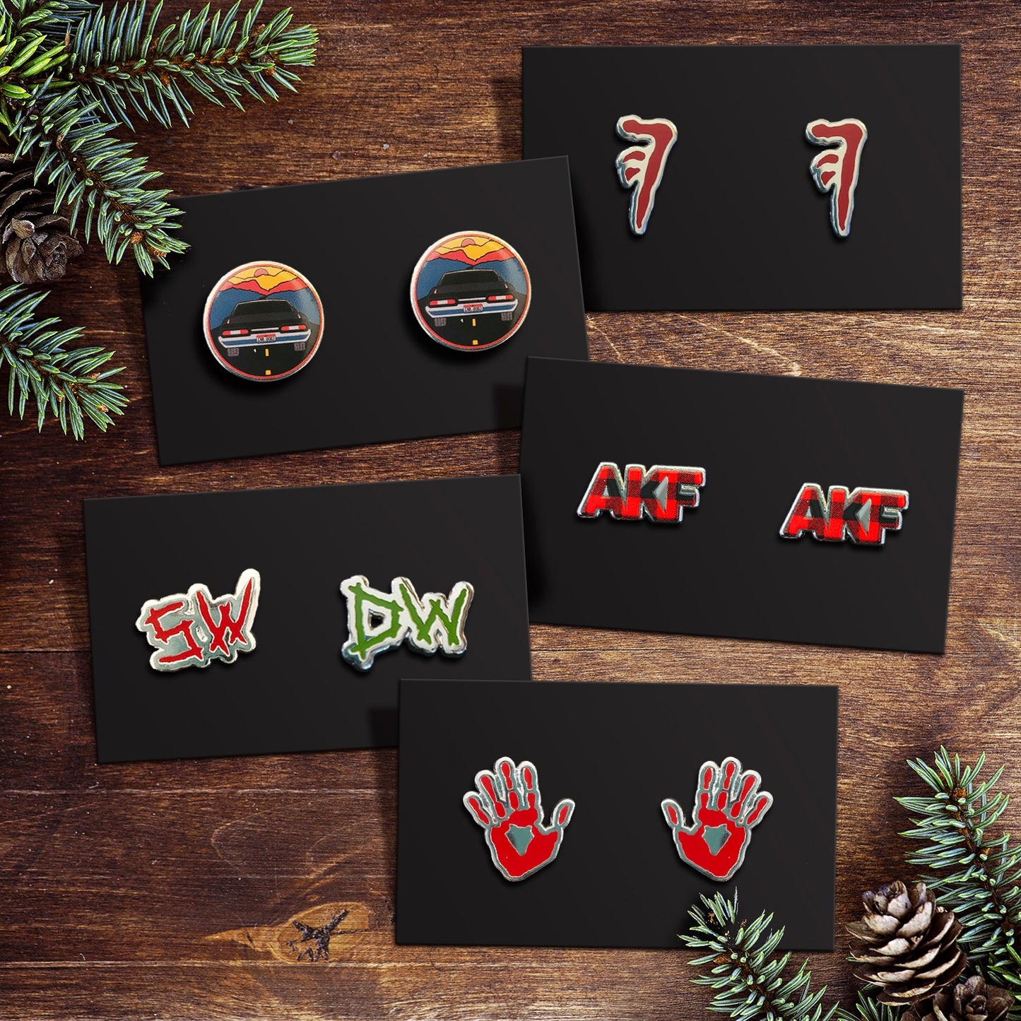 Five pairs of earrings, each on a black card, against a wooden table. One pair depicts red hands, one depicts the Mark of Cain, one depicts AKF in red and black plaid, one is the initial SW and DW in red and green, and one depicts a black chevy Impala diving toward a sunset.