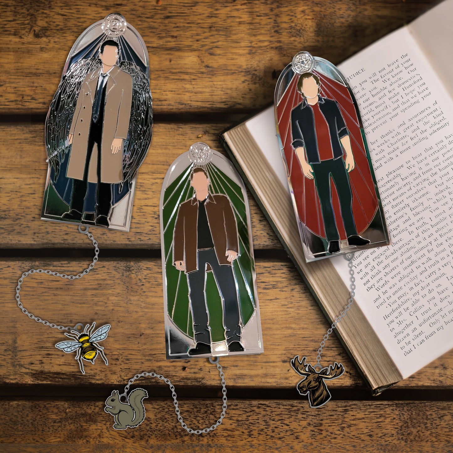 Three bookmarks on a wooden table. The left bookmark depicts Castiel against a blue background, the center has Dean Winchester against a green background, the right has Sam Winchester against a red background. Each bookmark has a charm attached to it by a chain at the bottom. Castiel's is a bee, Dean's is a Squirrel, Sam's is a Moose. An open book is under the Sam bookmark.