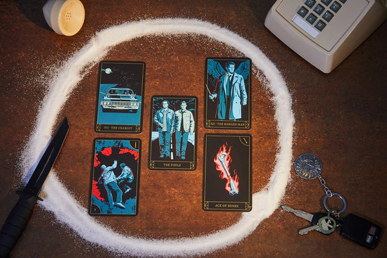 Five tarot cards depicting characters from the TV series Supernatural on a brown table. The cards are surrounded by a circle of salt. Around the salt are an old telephone, a hunting knife, and a set of car keys with an anti-possession symbol keychain.