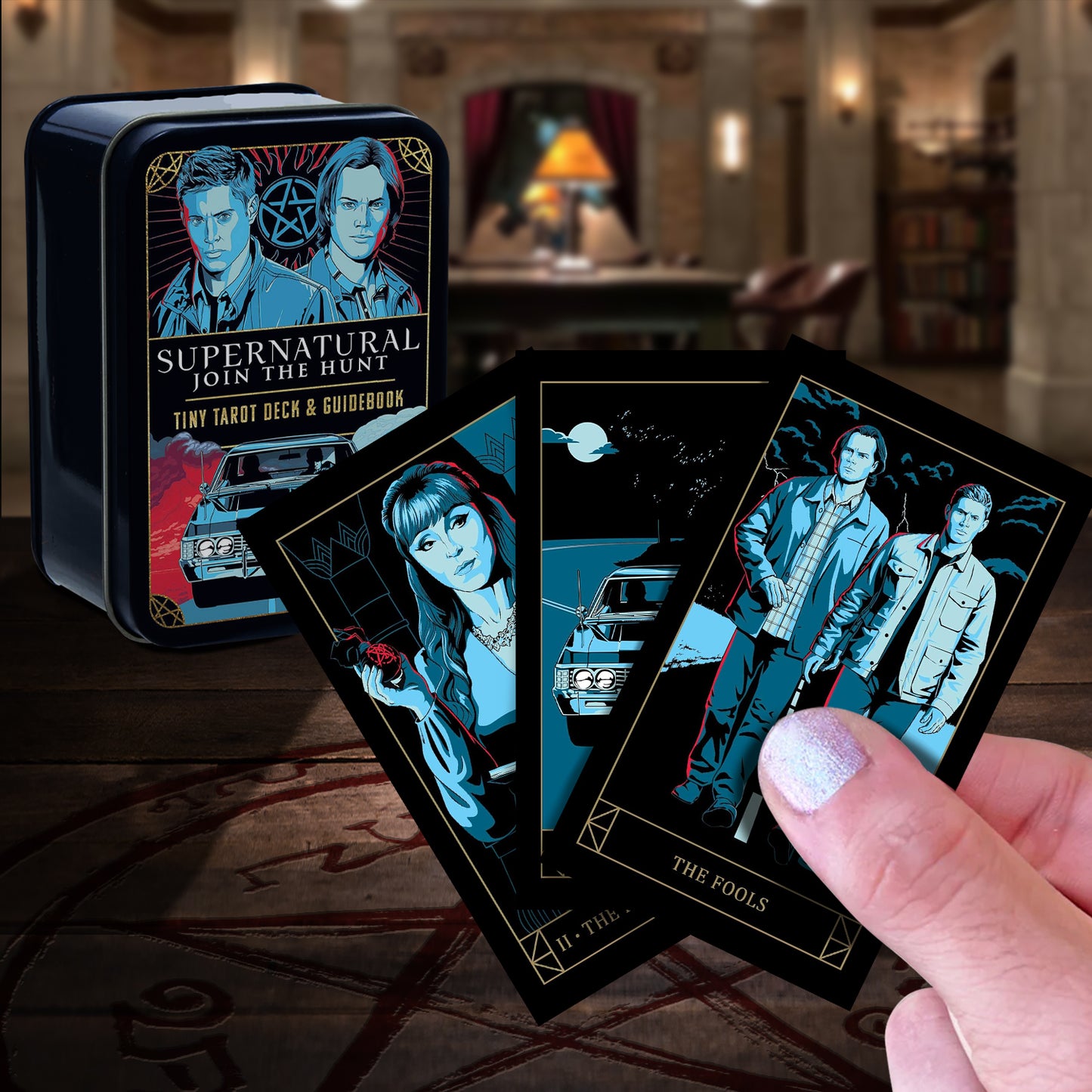 Supernatural-themed tarot cards with a tin box on a wooden surface. In the background is The Bunker. A model's hand is holding three of the cards in the foreground.