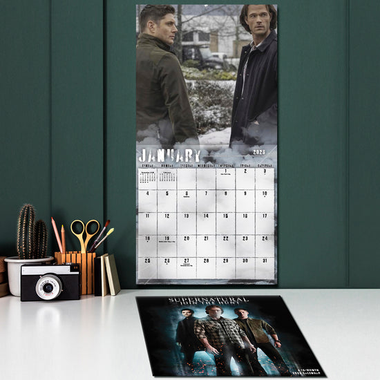 Supernatural-themed calendar on a desk with office supplies against a green wall.
