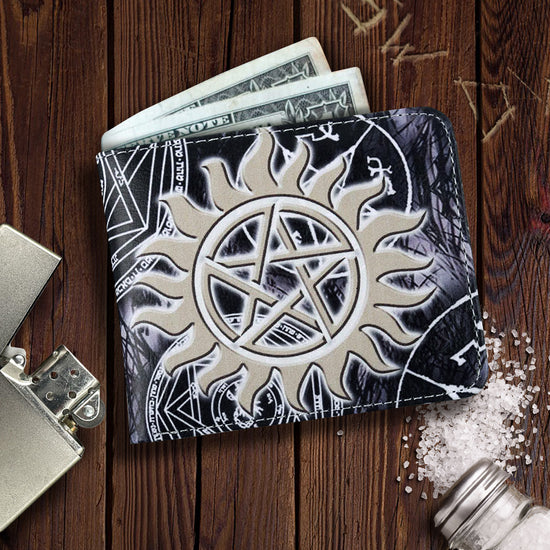 A black bifold wallet with the "Supernatural" logo printed on the back, a large white anti-possession symbol printed on the front, and an all-over print of angel sigils and devil's traps. The wallet is resting on a wooden table with "SW" and "DW" carved into it and there is a lighter and spilled salt shaker nearby.