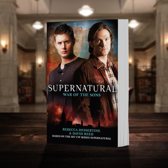 A copy of the book "Supernatural: War of the Sons" pictured in the "bunker" from Supernatural. The cover shows early-season Sam and Dean Winchester and reads "Supernatural War of the Sons - Based on the hit CW series SUPERNATURAL by Rebecca Desertine and David Reed”