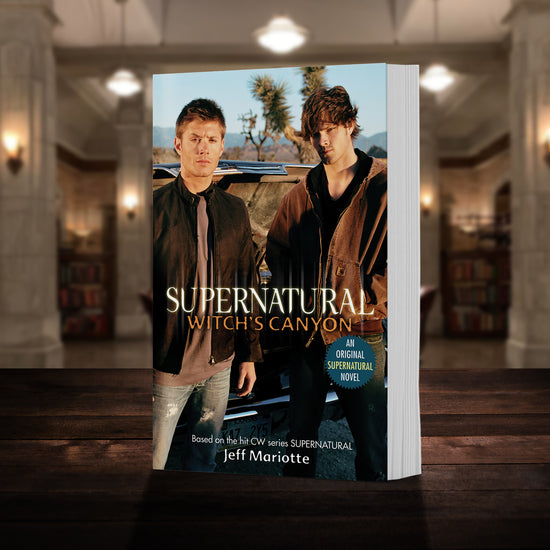 Load image into Gallery viewer, A copy of the book &amp;quot;Supernatural: Witch&amp;#39;s Canyon&amp;quot; pictured in the &amp;quot;bunker&amp;quot; from Supernatural. The cover shows early-season Sam and Dean Winchester and reads &amp;quot;Supernatural Witch&amp;#39;s Canyon - Based on the hit CW series SUPERNATURAL by Jeff Mariotte&amp;quot;
