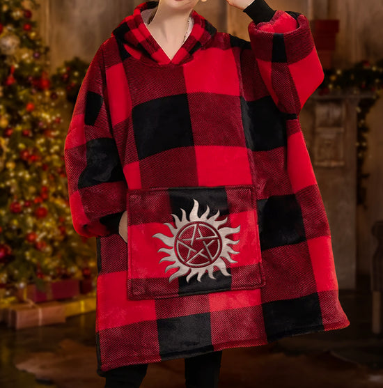 A female model wearing an oversized hoodie sweatshirt. The sweatshirt has a black and red plaid pattern, and is shaped like a wearable blanket. In the background is a Christmas tree with gifts underneath it, against a marble wall.