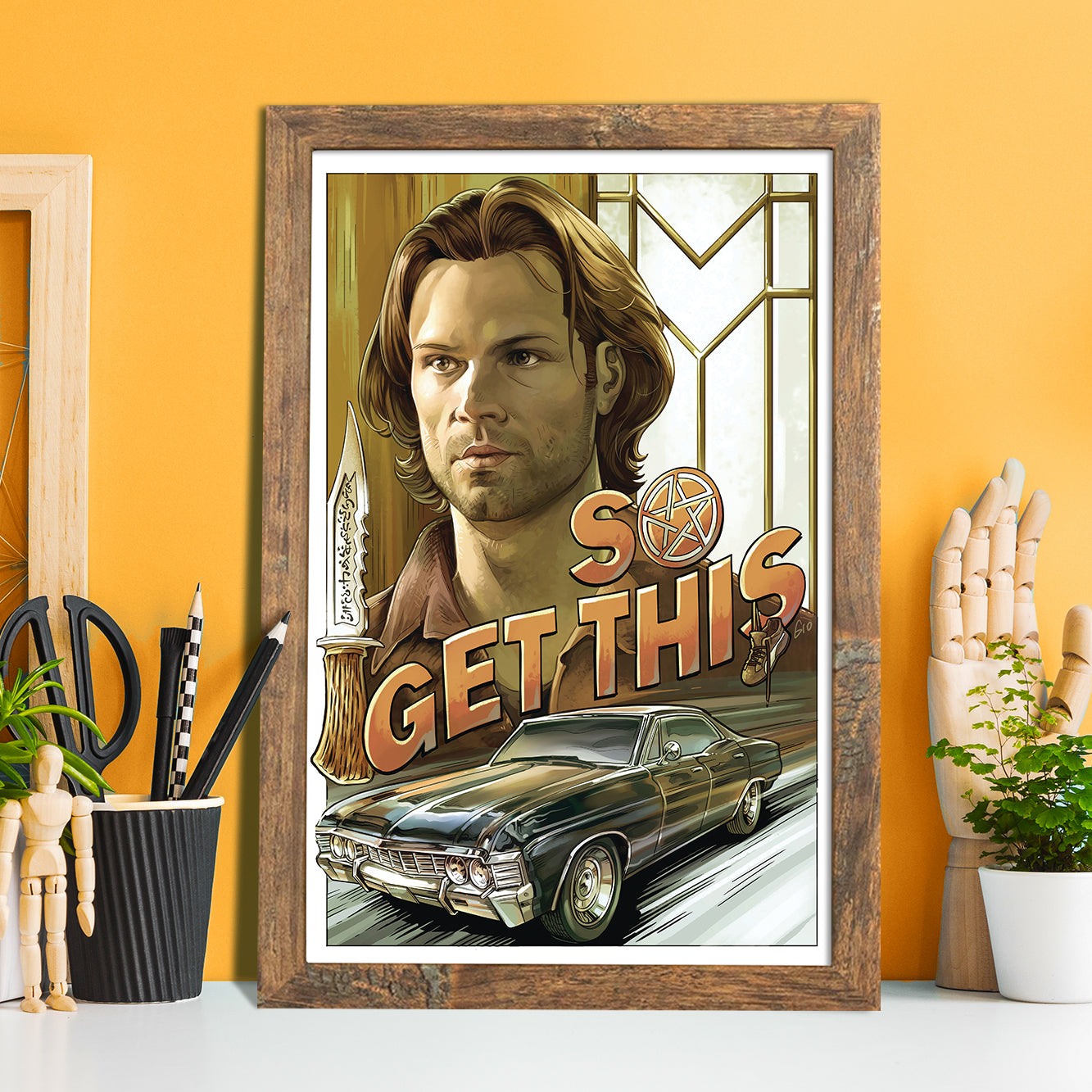 A framed drawing of Sam Winchester's face. Under the face is a 1967 Chevy Impala, with orand text above it saying "so get this." The O in the text contains the anti-possession symbol. Behind the drawing is a yellow wall. On either side are artist's tool and wooden models.