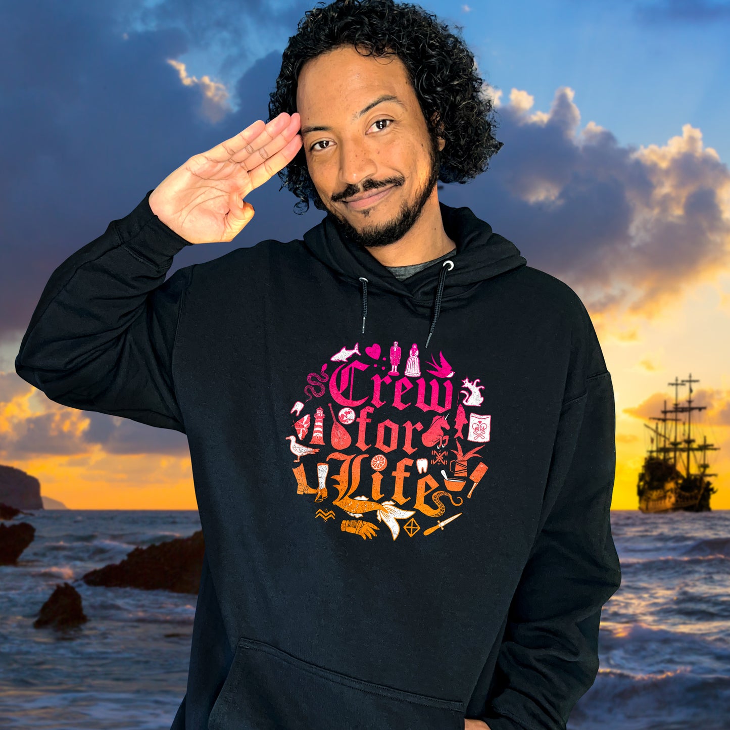 An image of actor Samba Schutte in a black hoodie. On the front of the shirt is pink and orange old fashioned text that says "Crew for life." Around the text are drawings of flowers, gloves, and chef tools. Behind Samba is a pirate ship on the open water, under a blue and yellow sky.