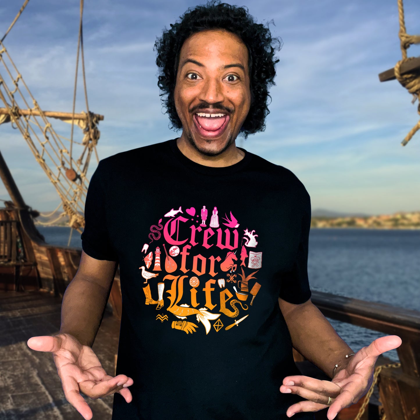 An image of actor Samba Schutte in a black T-shirt. On the front of the shirt is pink and orange old fashioned text that says "Crew for life." Around the text are drawings of flowers, gloves, and chef tools. Behind Samba is a pirate ship on the open water, with land in the distance.