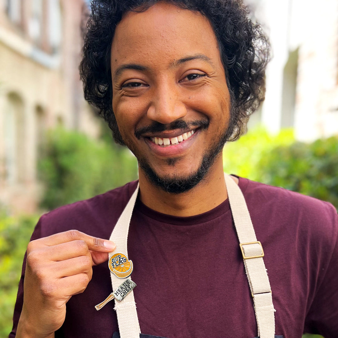 A waist-up image of actor Samba Schutte wearing a burgandy t-shirt. Cream apron straps are visible, and on the straps are set of two colored enamel pins: an orange slice with "our flag" in writing and a meat cleaver with "means chef" in writing.