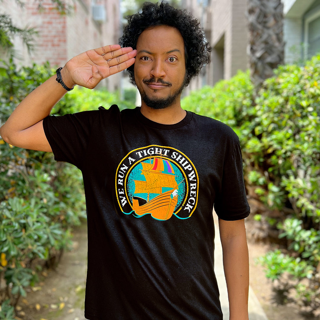 Actor Samba Schuttel wearing a black T-shirt and standing in front of greenery. On the shirt are white letters saying "We Run A Tight Shipwreck" are in an arc around the middle, with a pirate ship inside the arc. The pirate ship's flag is in rainbow colors.