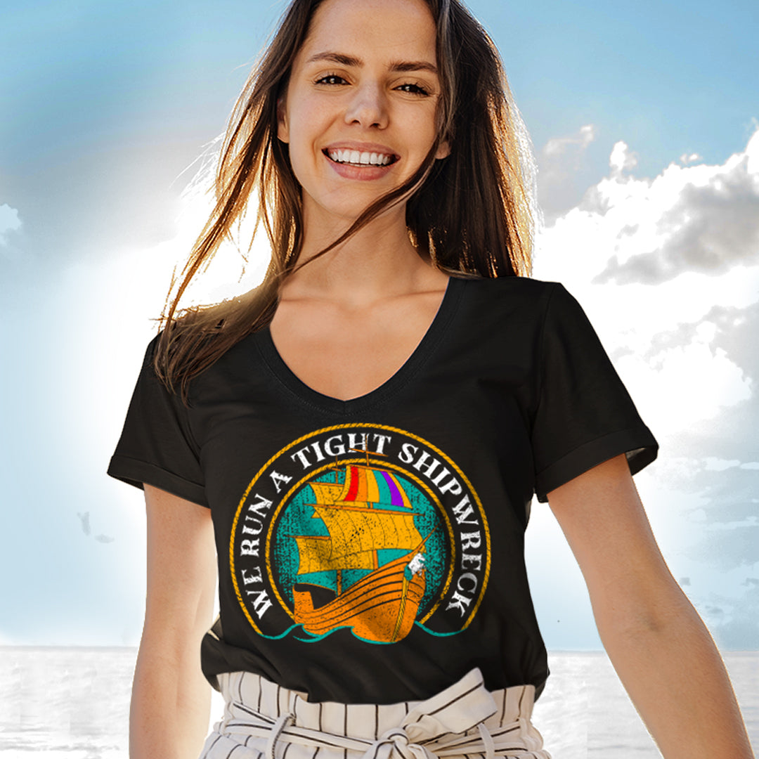 A female model wearing a black T-shirt, standing in front of a blue sky on the beach. On the shirt are white letters saying "We Run A Tight Shipwreck" are in an arc around the middle, with a pirate ship inside the arc. The pirate ship's flag is in rainbow colors.
