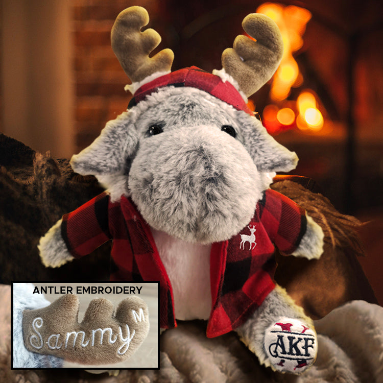 Plush moose toy with antlers and a red plaid shirt, sitting in front of a fireplace. Inset is a close up of one antler, with "Sammy" stitch on it in white thread.
