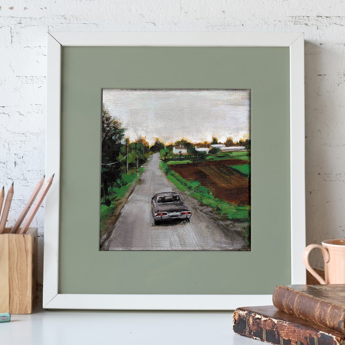 A white-framed lithograph against a white stone wall. The lithograph depicts a black Impala driving down a dirt road next to a farm. In front of the lithograph is a pencil holder and a pair of leather-bound books.