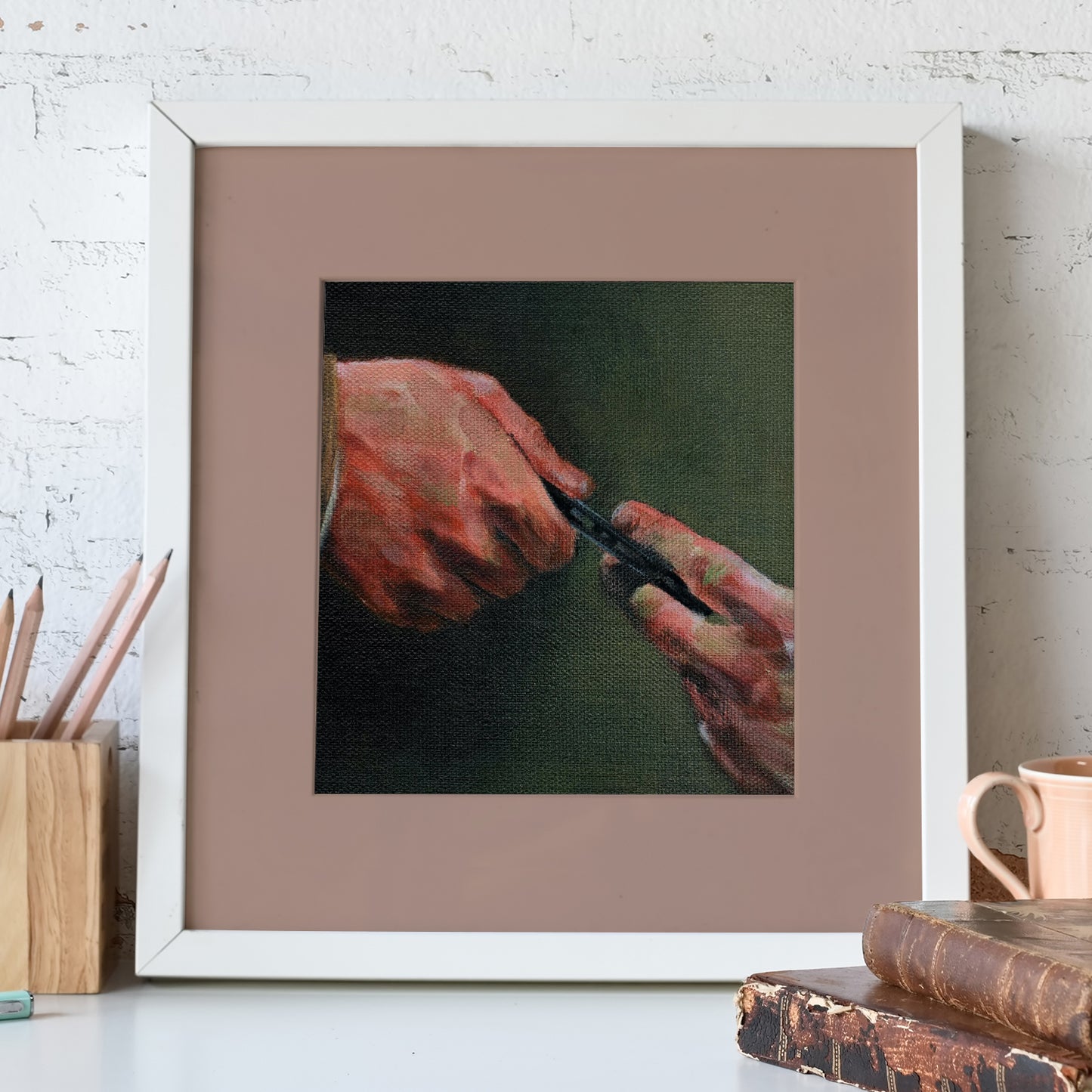 A white-framed lithograph against a white stone wall. The lithograph depicts a pair of hands against a green background. One hand is clutching a black cassette tape, giving it to the other In front of the lithograph is a pencil holder and a pair of leather-bound books.