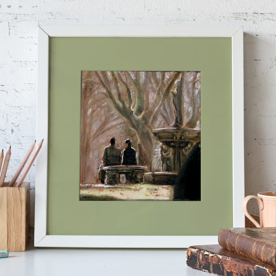 A white-framed lithograph against a white stone wall. The lithograph depicts two SPN characters sitting on a stone bench in a park, next to a stone fountain. In front of the lithograph is a pencil holder and a pair of leather-bound books.