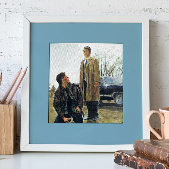 A painting mounted on a blue background in a white frame. The painting shows Dean Winchester and the angel Castiel in a field next to the Impala. Behind the painting is a white brick wall.