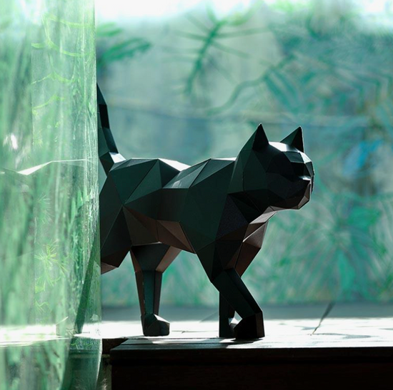 A paper model of a black cat is standing on a window sill, partially covered by a green curtain. Behind the cat are tree branches.